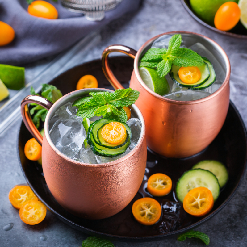 Two copper mugs of Cucumber Mint Moscow Mule, decorated with cucumber slices and mint leaves on a serving plate.