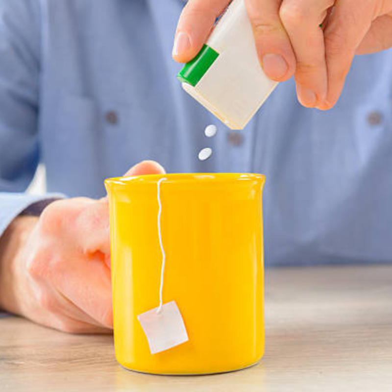 A person adds sugar to a yellow cup, demonstrating the use of artificial sweeteners in beverage preparation.